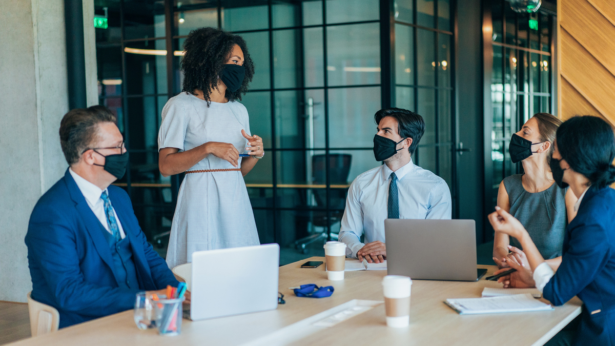 A Black woman stands to speak to fellow board members seated around a table