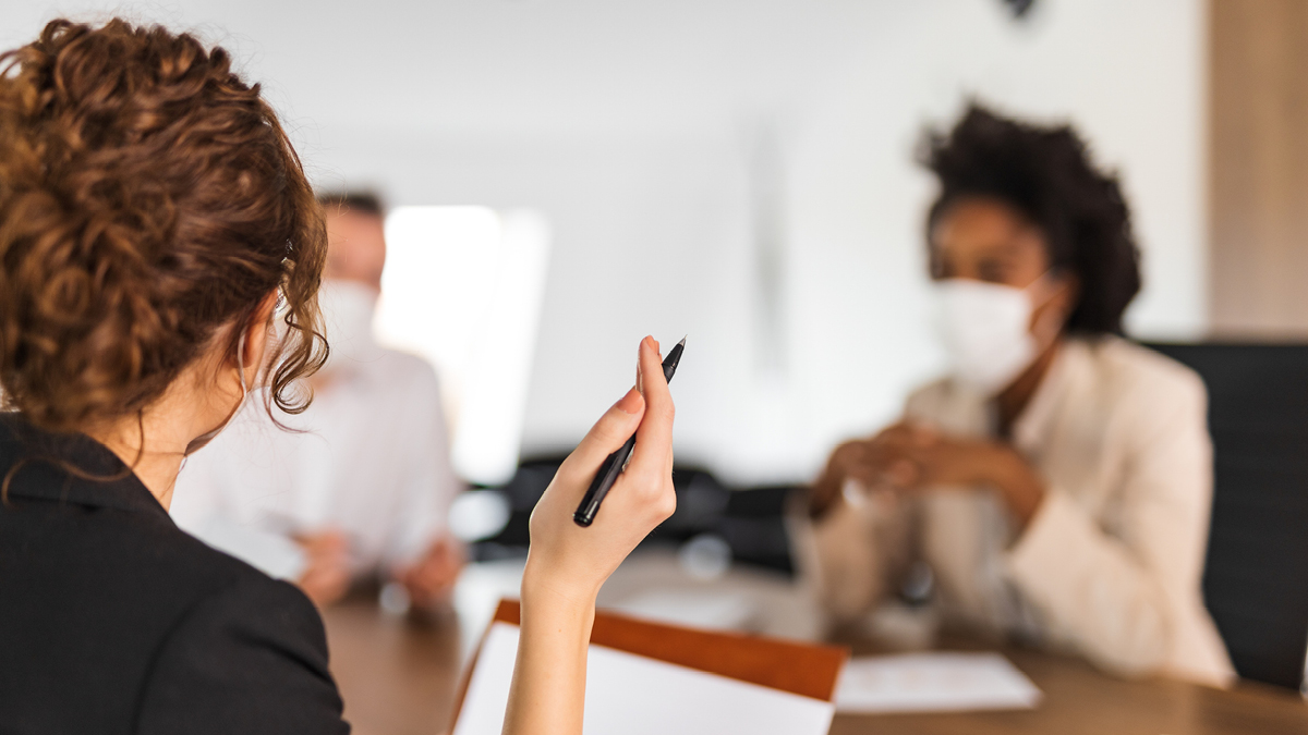 A white woman speaks to two other board members, a white man and a Black woman, seated at a table