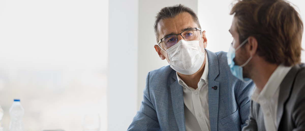 Two men, both masked, sit at a table and discuss
