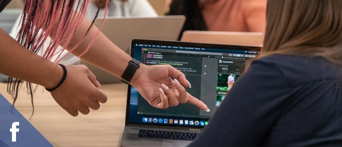 Close up image of a woman (standing) pointing and instructing another woman (seated) on how to do something on a laptop.