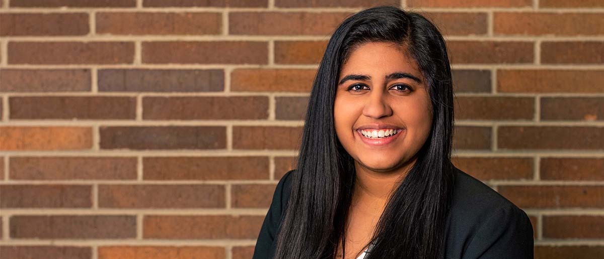 A woman with long dark hair wears a professional blazer and poses in front of a brick wall 