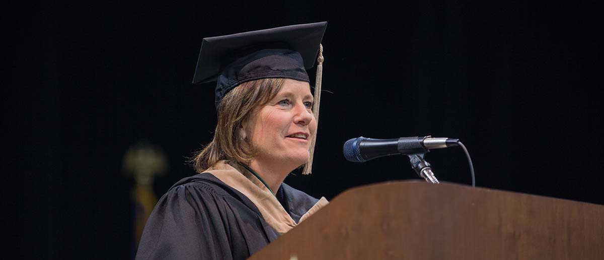 Linda Hubbard wears her black commencement ceremony cap and gown as she stands at the podium and addresses the graduating students