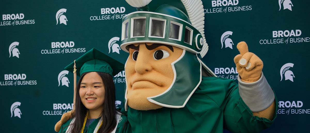 A Broad College student takes a picture with the mascot Sparty in front of Broad College of Business backdrop