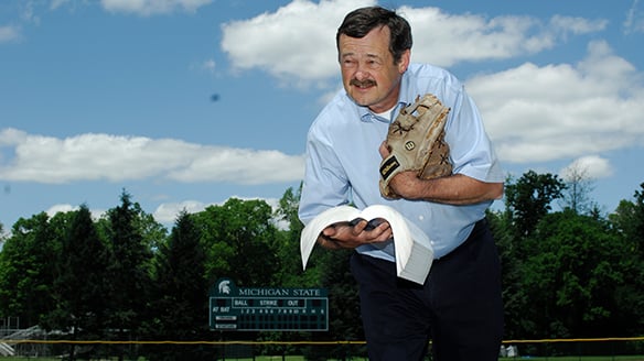 Ed Outslay hold a book in one hand and wears a baseball glove on the other with a clear, blue sky behind him
