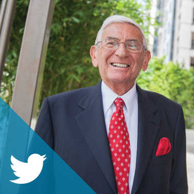 Eli Broad in a navy suit and red tie stands outside on a sunny day