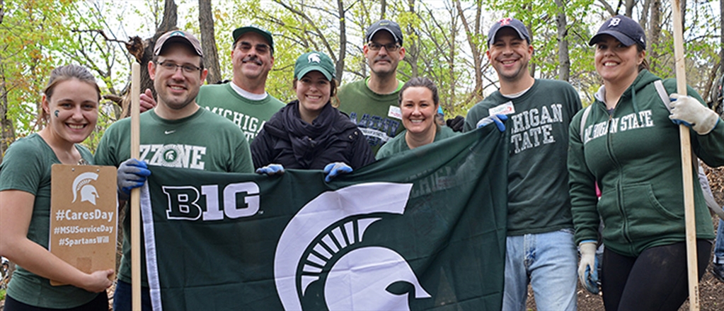 Alumni pose with a Spartan flag at their service day in the woods