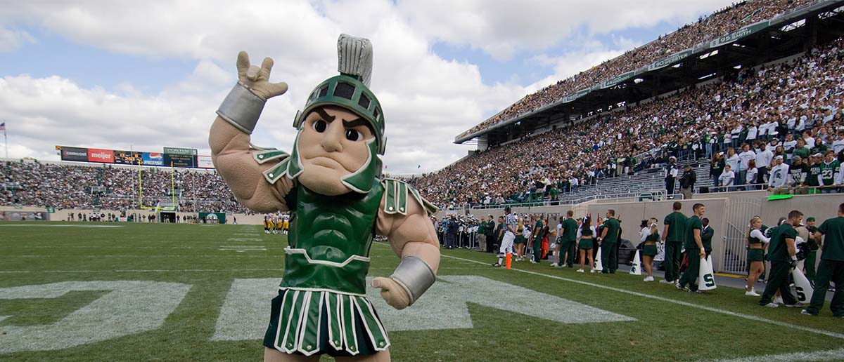 Sparty stands on the football field and signs "I love you" in American Sign Language.