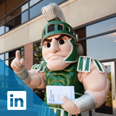Sparty stands outside the home of the Gast Business Library holding a copy of the Accounting Review