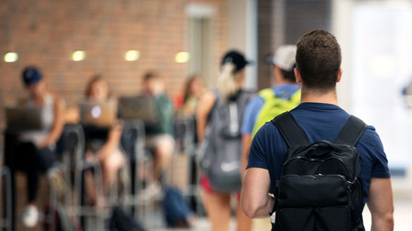 Male student with backpack walks down BCC hallway past students on computers at side tables