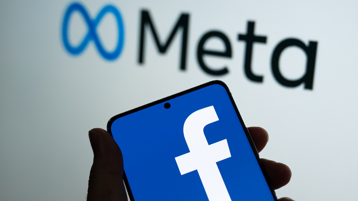 Facebook logo on a phone held in front of Meta logo on a wall