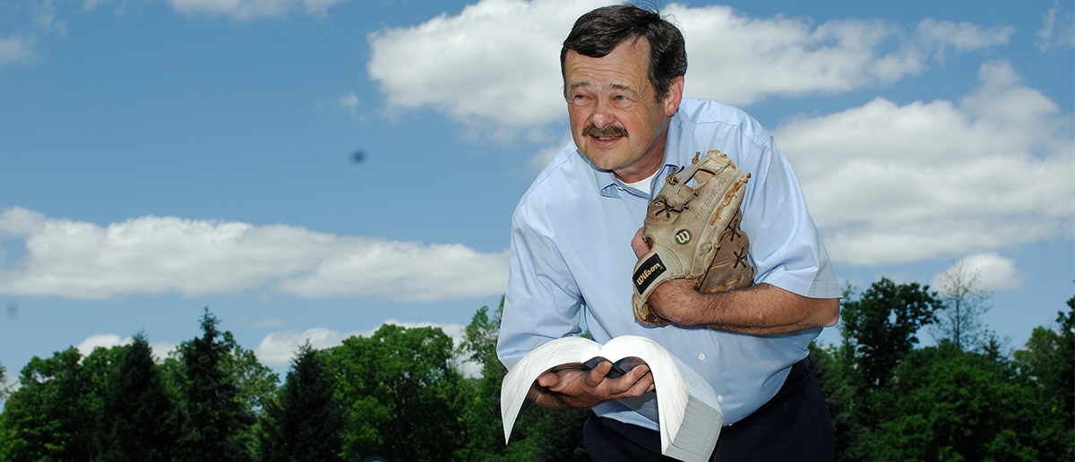 Ed Outslay hold a book in one hand and wears a baseball glove on the other with a clear, blue sky behind him