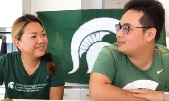 Broad alumni Yvonne Lo and Calvin Lai represent MSU with green and white shirts and a flag for their virtual cooking class event.